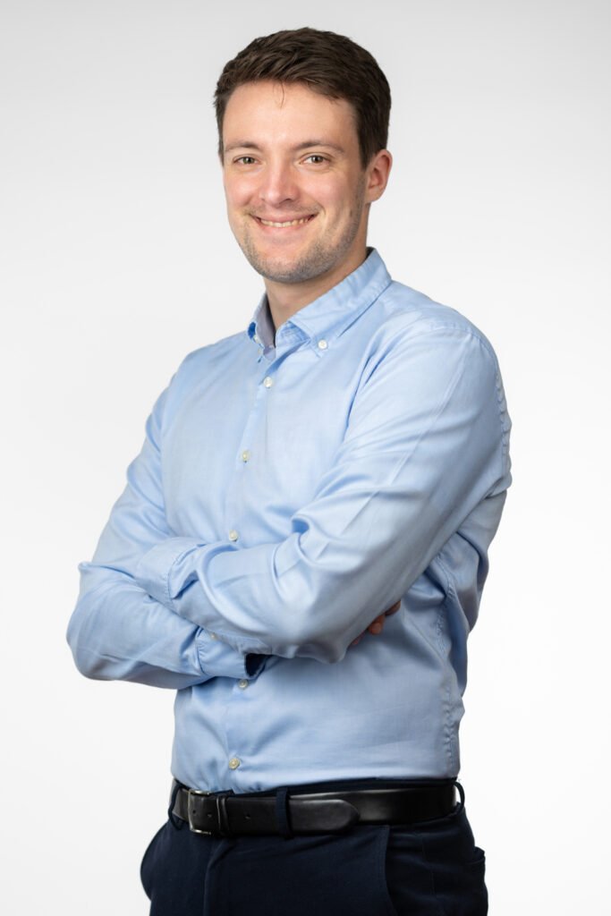 A man, sustainability consultant Hannes Lefherz, stands with his arms folded and wears a light blue button-down shirt. He combines the shirt with dark trousers and a black belt. The background is plain and white, which draws the focus to the person.