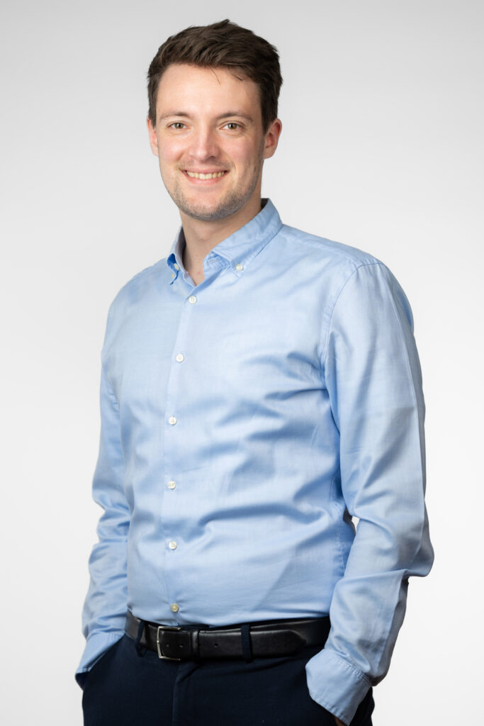 Ein Mann, Nachhaltigkeitsberater Hannes Lefherz, trägt ein hellblaues Hemd mit Knöpfen und hat die Ärmel leicht aufgekrempelt. Er kombiniert das Hemd mit einer dunklen Hose und einem schwarzen Gürtel. Der Hintergrund ist schlicht und weiß, was den Fokus auf die Person lenkt.