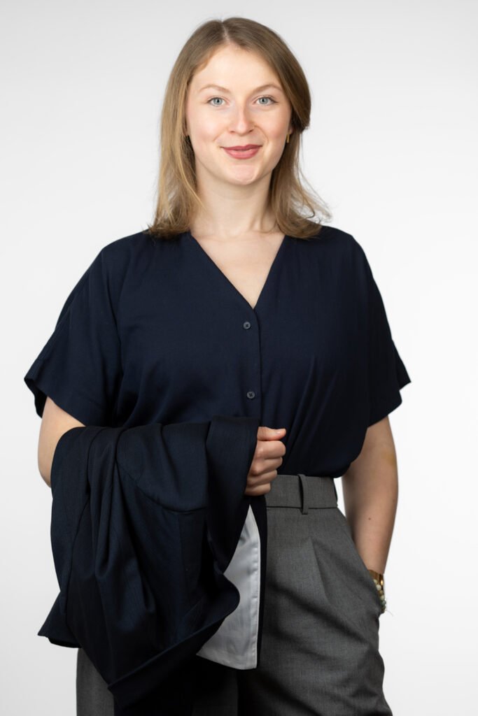 A woman, sustainability consultant Lara Morlang, wears a dark blue blouse with a V-neck and has a dark blue blazer over her arm. She combines the blouse with gray pants. The background is plain and white, which puts the person in the foreground.