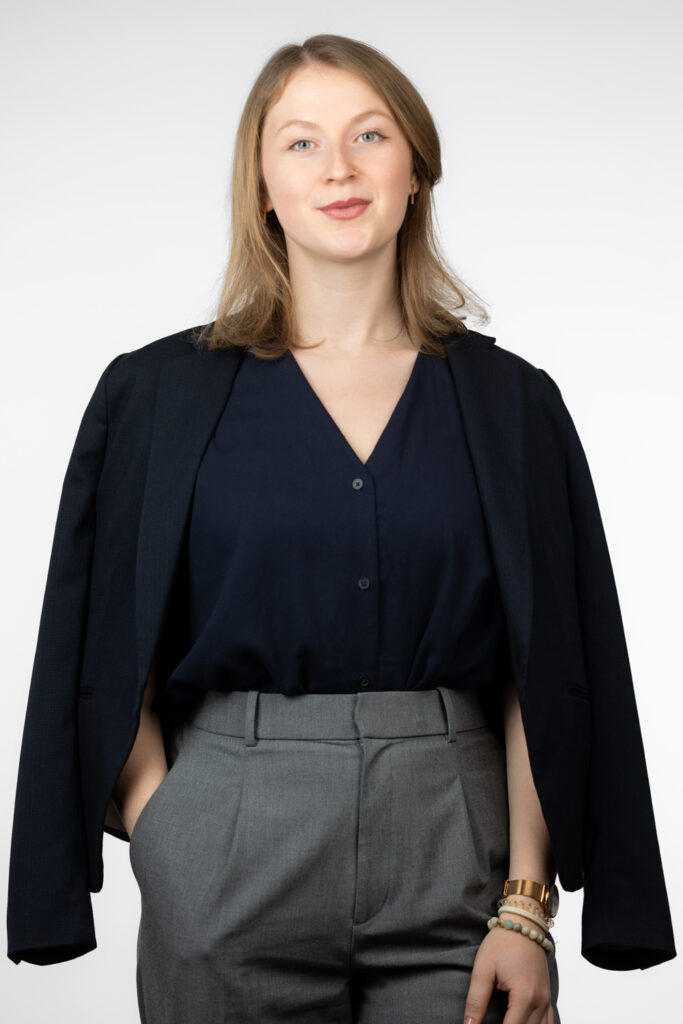 Eine Frau, die Nachhaltigkeitsberaterin Lara Morlang, trägt einen dunkelblauen Blazer über einer blauen Bluse und grauen Hosen. Sie hat mehrere Armbänder und eine Uhr an ihrem Handgelenk. Der Hintergrund ist schlicht und weiß, was den Fokus auf die Person legt.