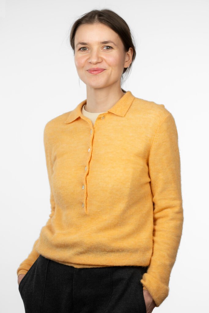 Eine Frau, Projektassistentin Sandrina Bahlo, trägt einen gelben Pullover mit Knöpfen und einem Kragen. Darunter ist ein helles Oberteil sichtbar. Sie kombiniert den Pullover mit dunklen Hosen. Der Hintergrund ist schlicht und weiß, was den Fokus auf die Person legt.