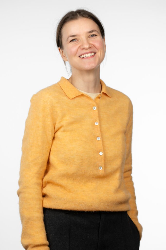 A woman, project assistant Sandrina Bahlo, wears a yellow sweater with buttons and a collar. The sweater has a soft and casual cut. A light-colored top is visible under the sweater. She combines the sweater with dark pants. The background is plain and white, which draws the focus to the person.